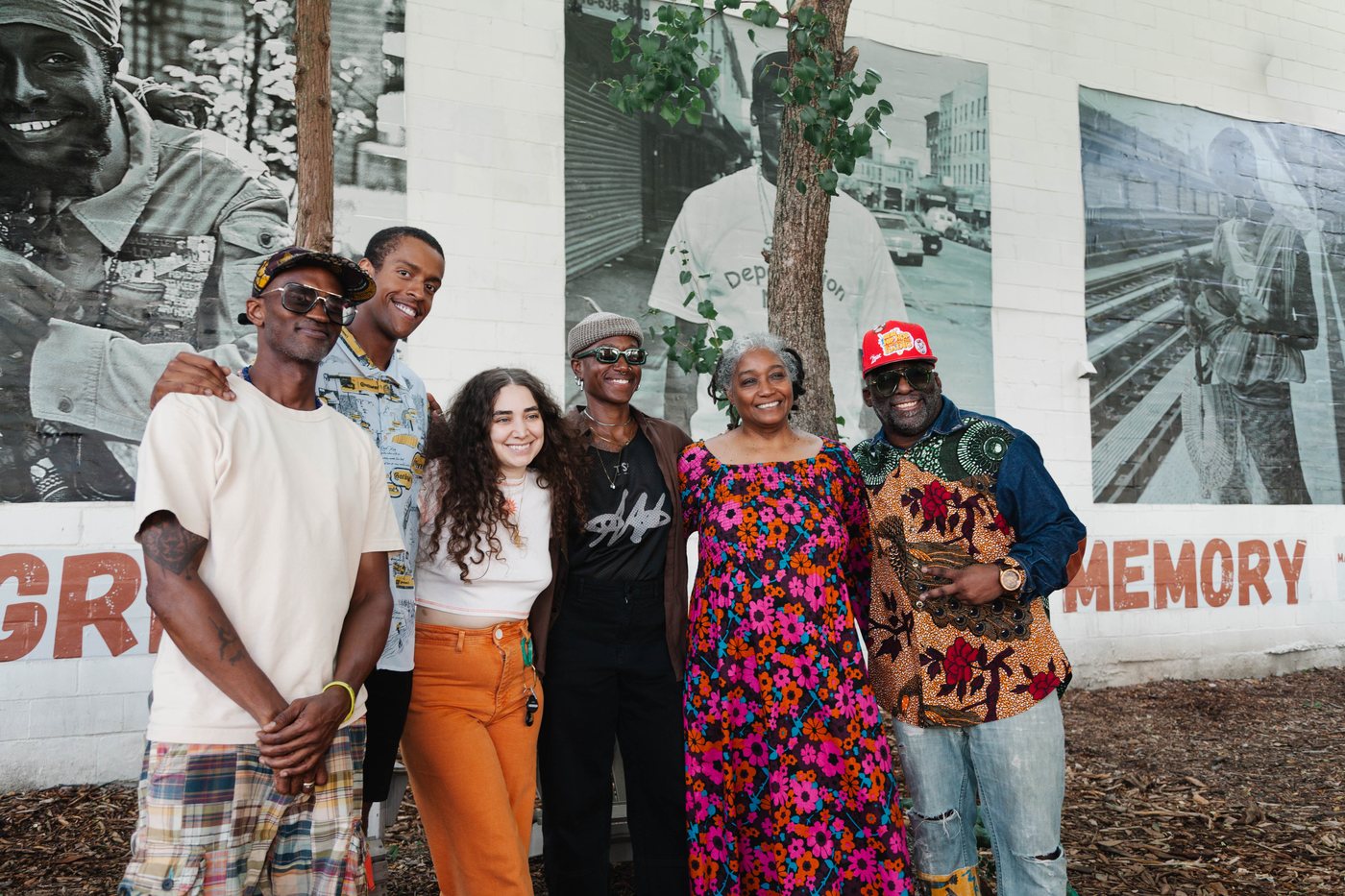 Six people stand arm in arm in front of the Griots of Visual Memory mural, smiling.