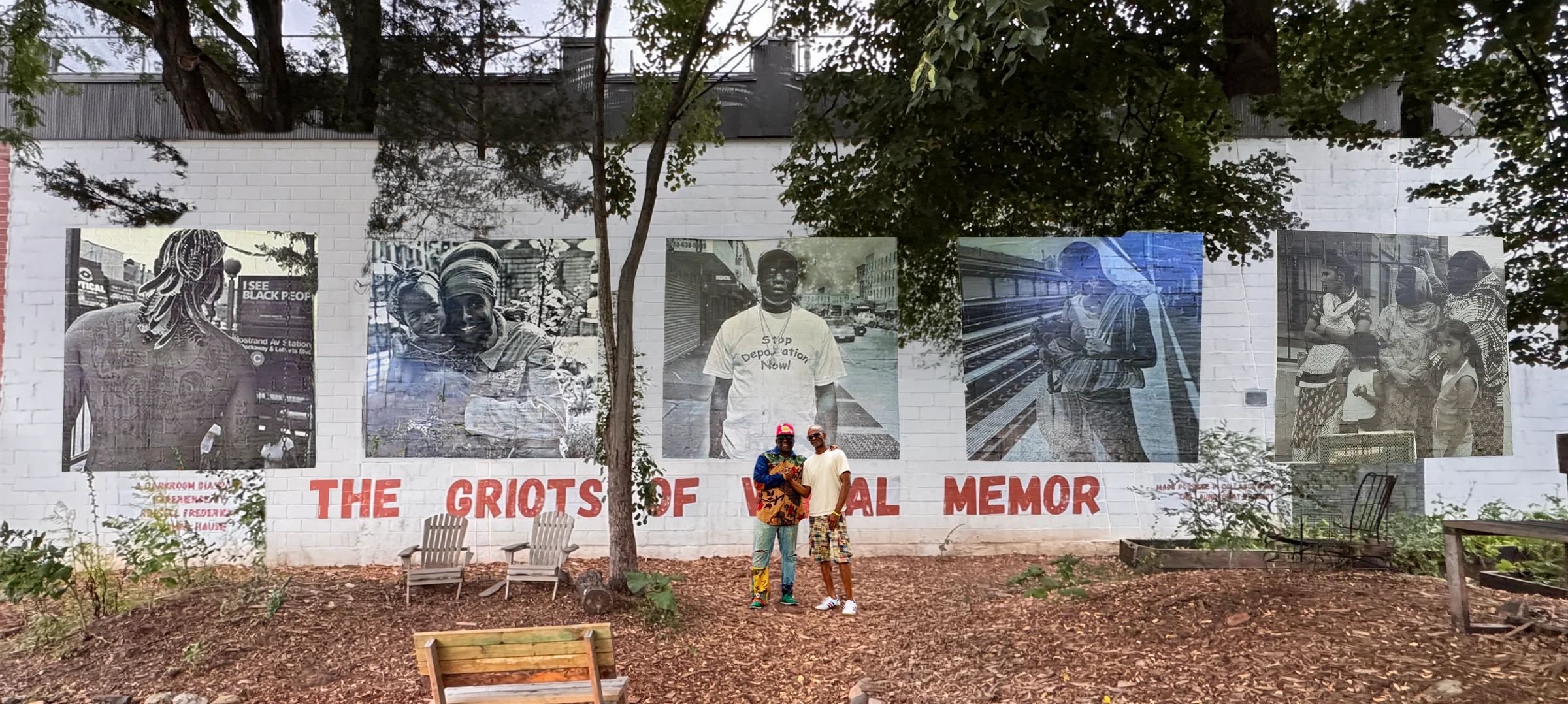 The Griots of Visual Memory mural at the Seeds of Somers Garden: five large black-and-white photographs wheatpasted on a white brick wall above the words THE GRIOTS OF VISUAL MEMORY painted in red. Russell Frederick and Dahkil Hausif stand in front of it.