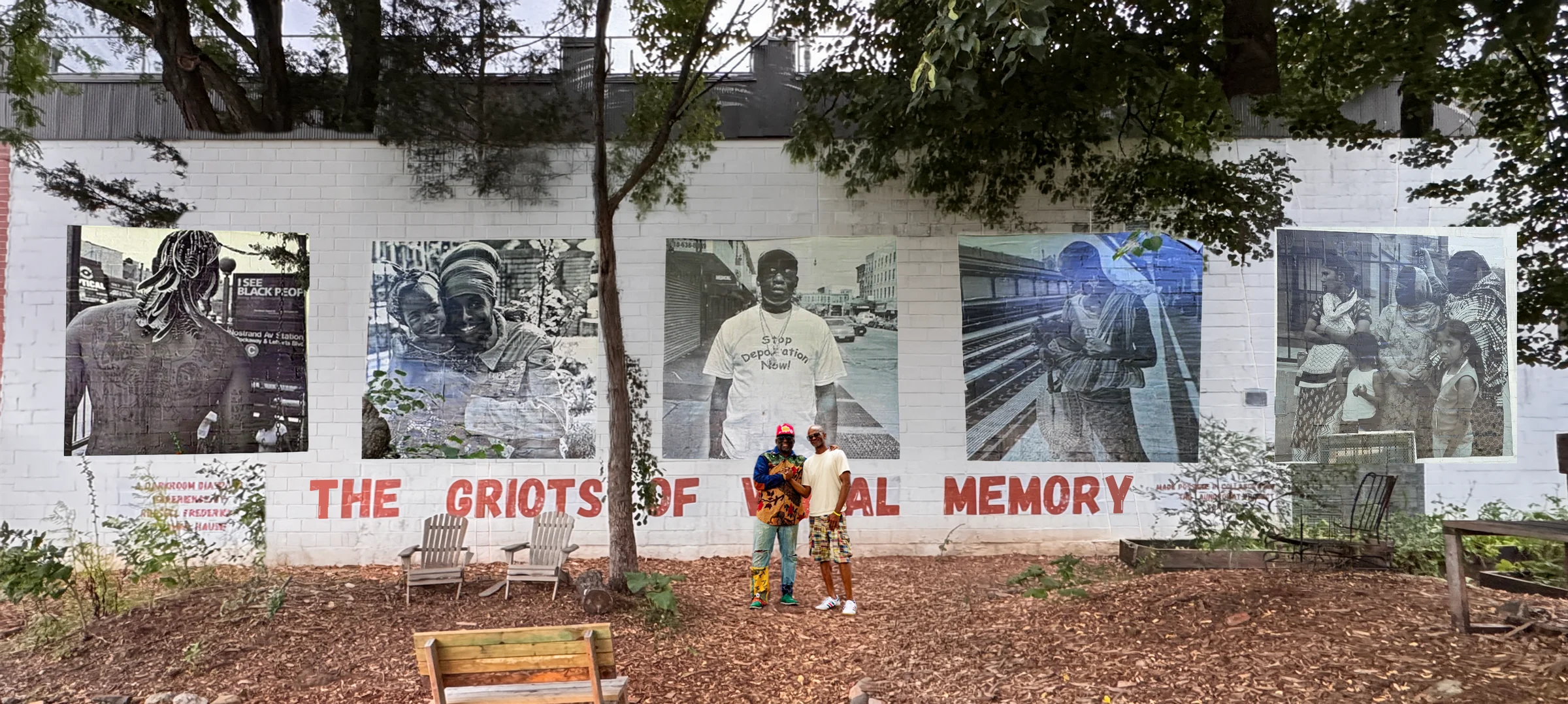 The Griots of Visual Memory mural at the Seeds of Somers Garden: five large black-and-white photographs wheatpasted on a white brick wall above the words THE GRIOTS OF VISUAL MEMORY painted in red. Russell Frederick and Dahkil Hausif stand in front of it.