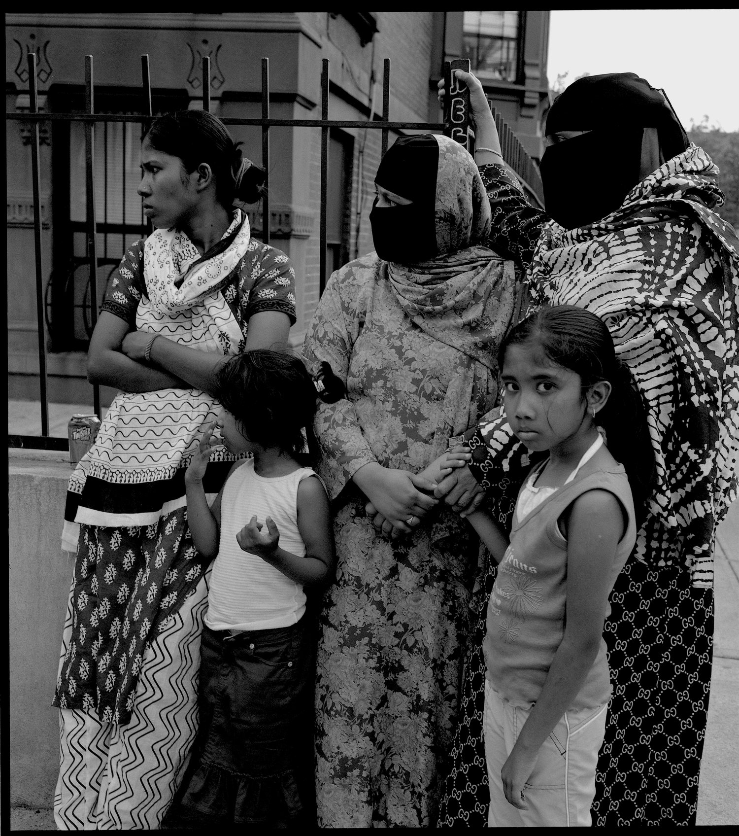 Three women and two children stand together on a Brooklyn sidewalk in front of an iron fence.