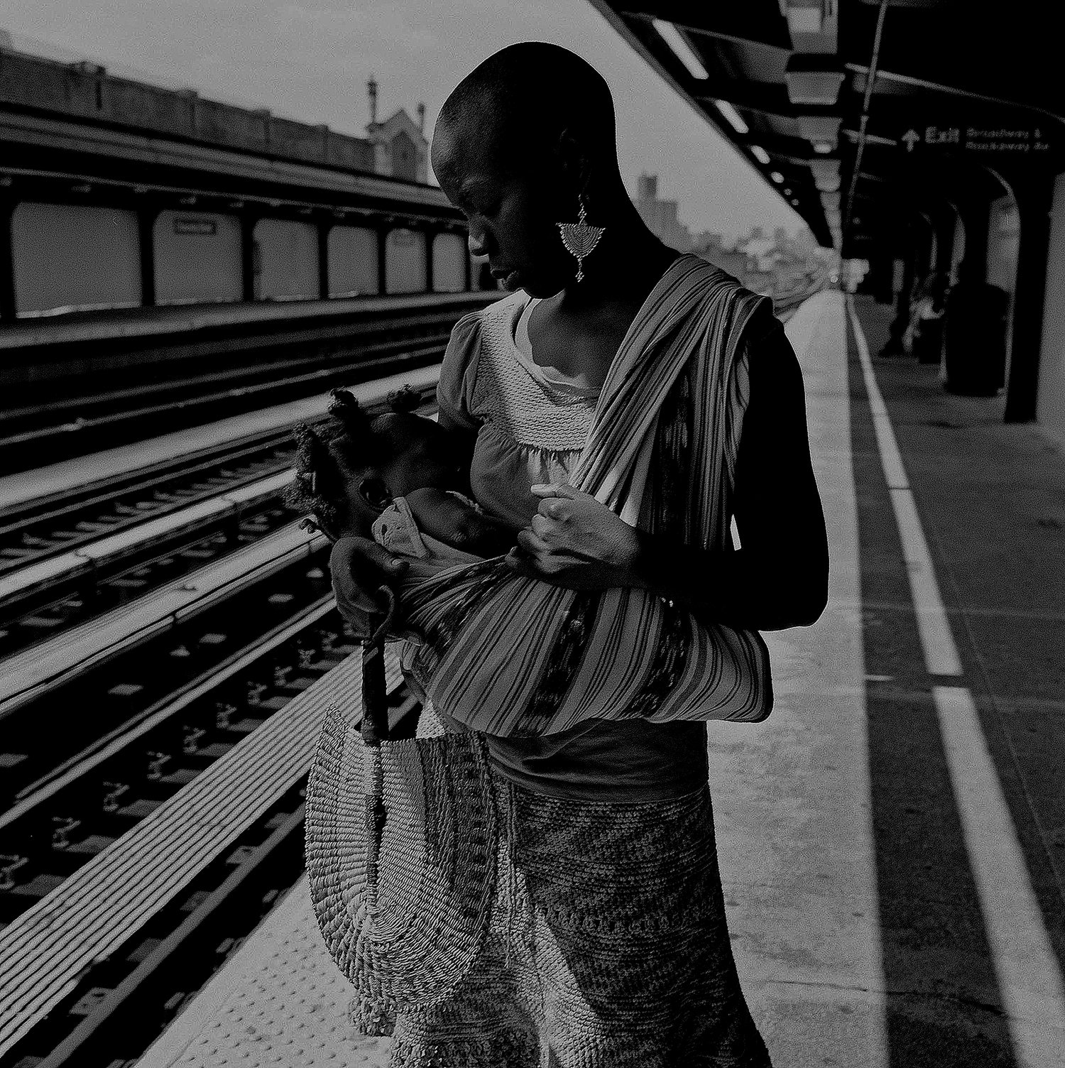 A woman stands on an elevated subway platform holding a sleeping baby in a striped wrap.