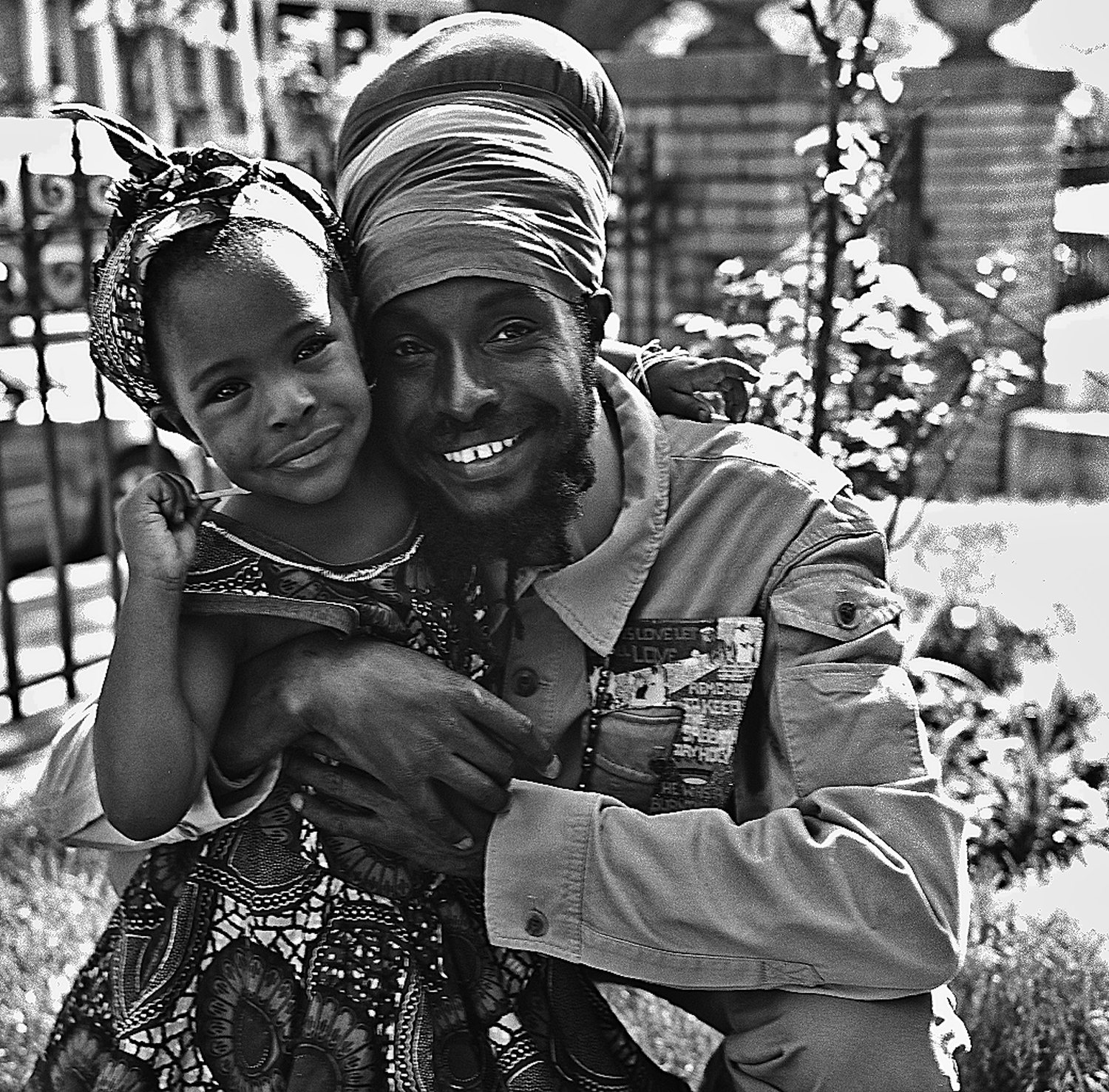A man in a headwrap holds a small child on his shoulder, both smiling, in a Brooklyn garden.