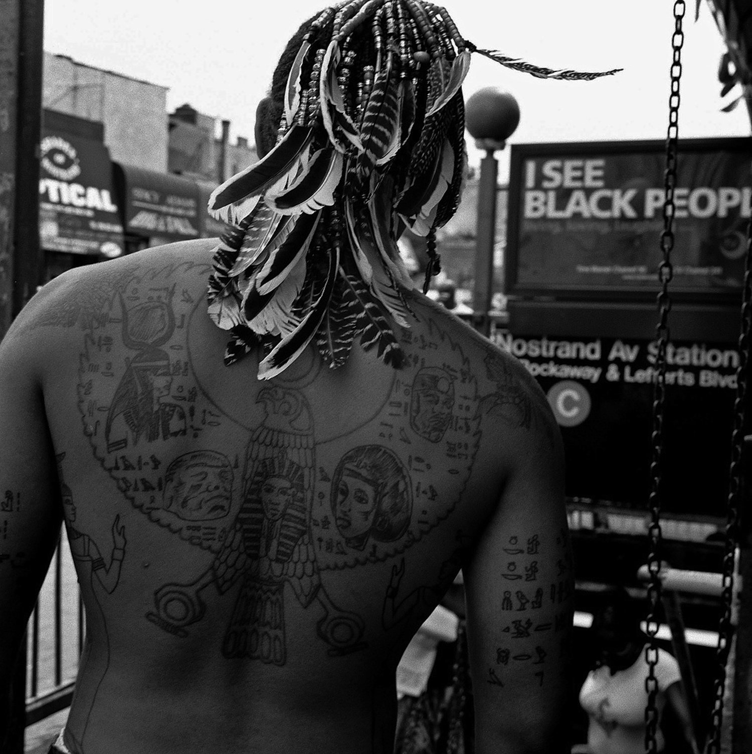 A man photographed from behind at Nostrand Avenue station, his back tattooed with hieroglyphs, feathers braided into his hair, beneath a sign reading I See Black People.